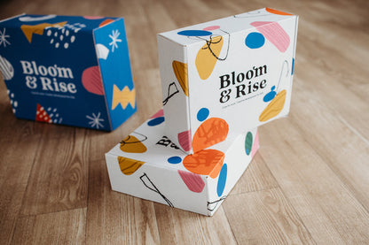 Three colorful Bloom & Rise gift boxes displayed on a wooden floor by Mama's Dream