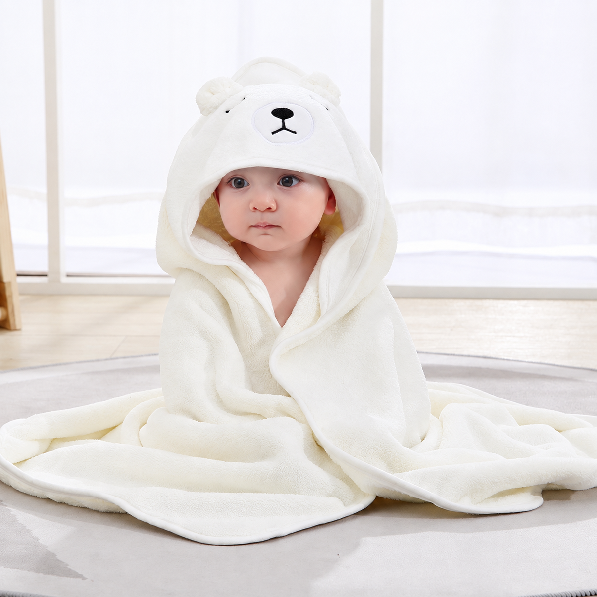 Baby sitting indoors wrapped in a soft cream hooded bath towel with an embroidered bear face by Mama's Dream