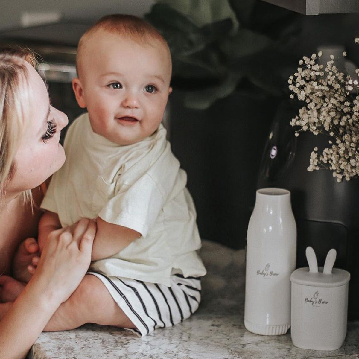 : Mother holding her baby beside a Baby's Brew portable bottle warmer and formula dispenser by Mama's Dream